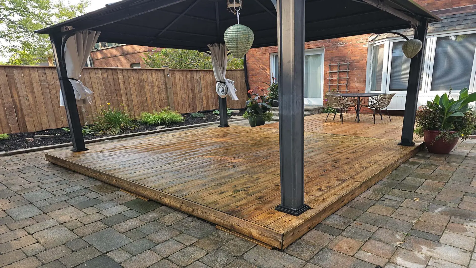 A freshly stained wooden deck with rich tones, under a gazebo in a cozy backyard space, surrounded by plants and outdoor furniture.