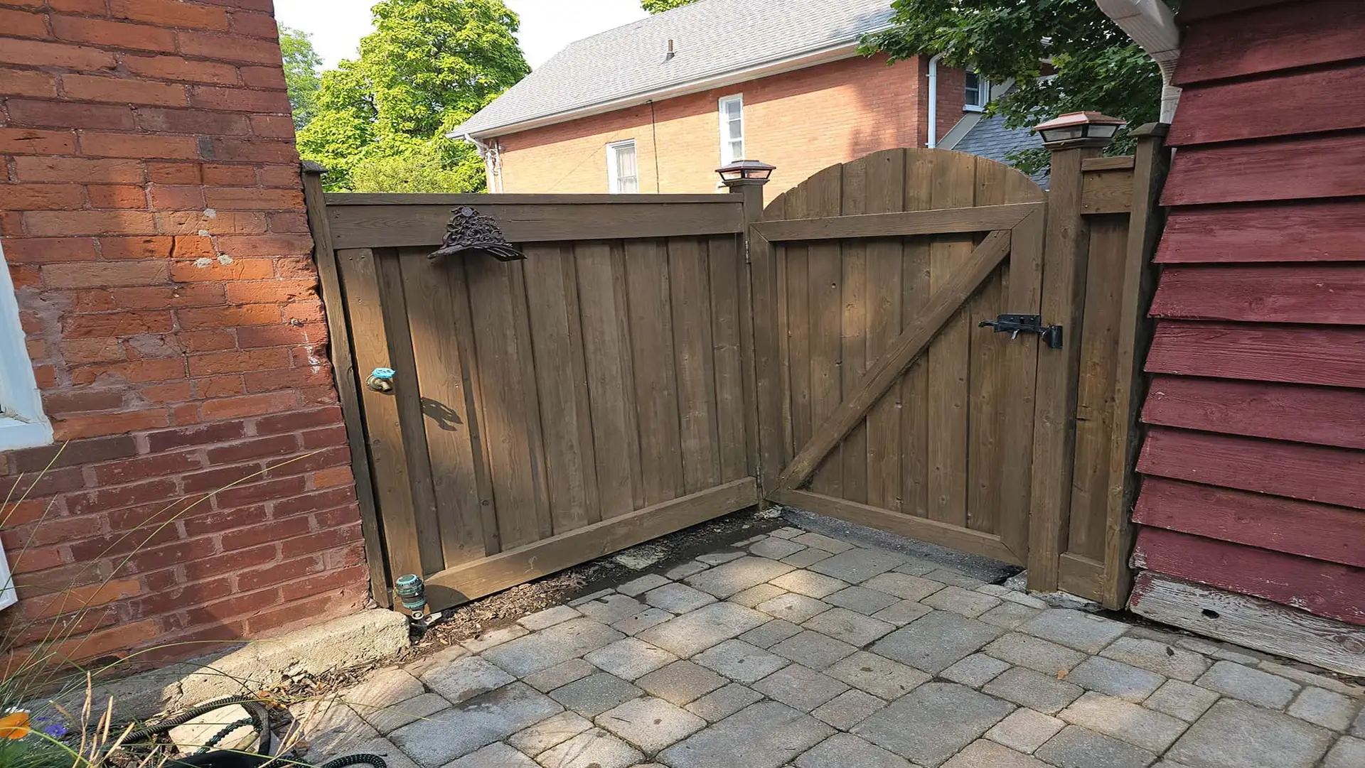 A wooden gate attached to a brick house, freshly stained with a dark finish, featuring a rustic metal latch and bell.