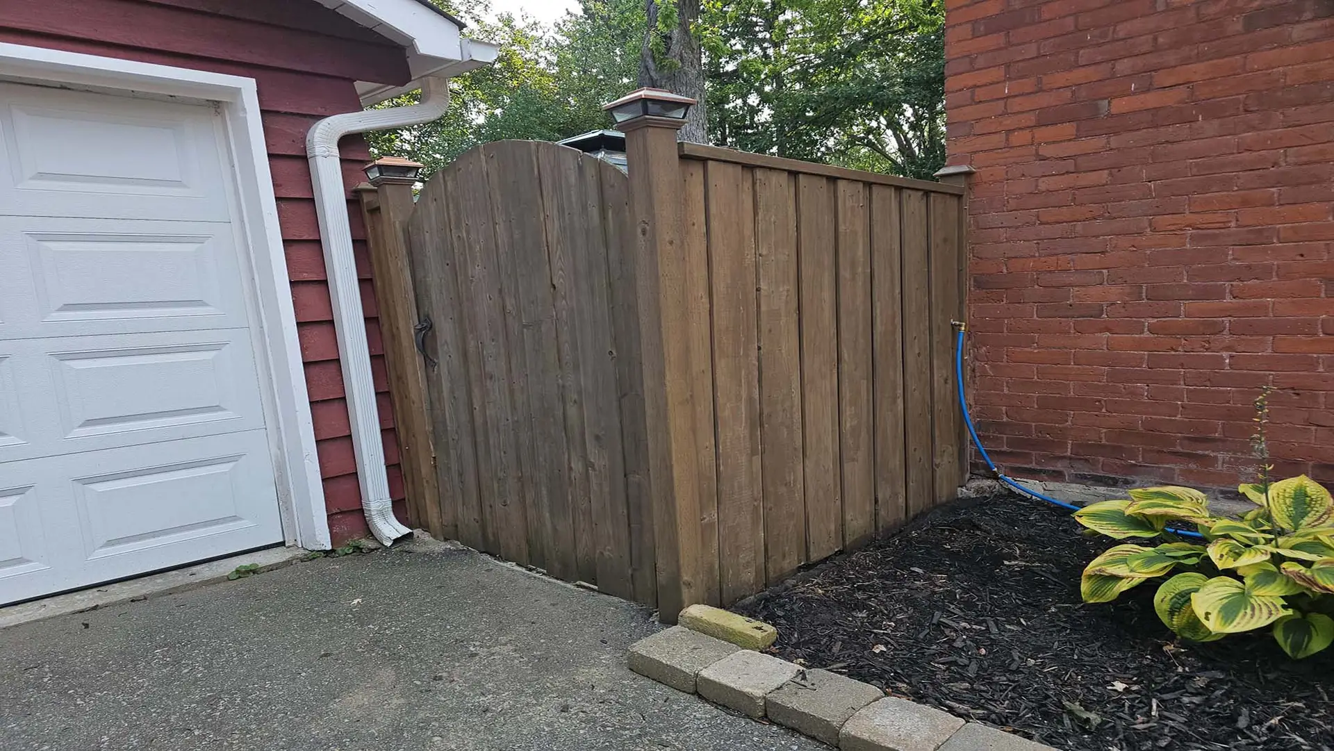 A wooden gate attached to a brick house, freshly stained with a dark finish, featuring a rustic metal latch and bell.